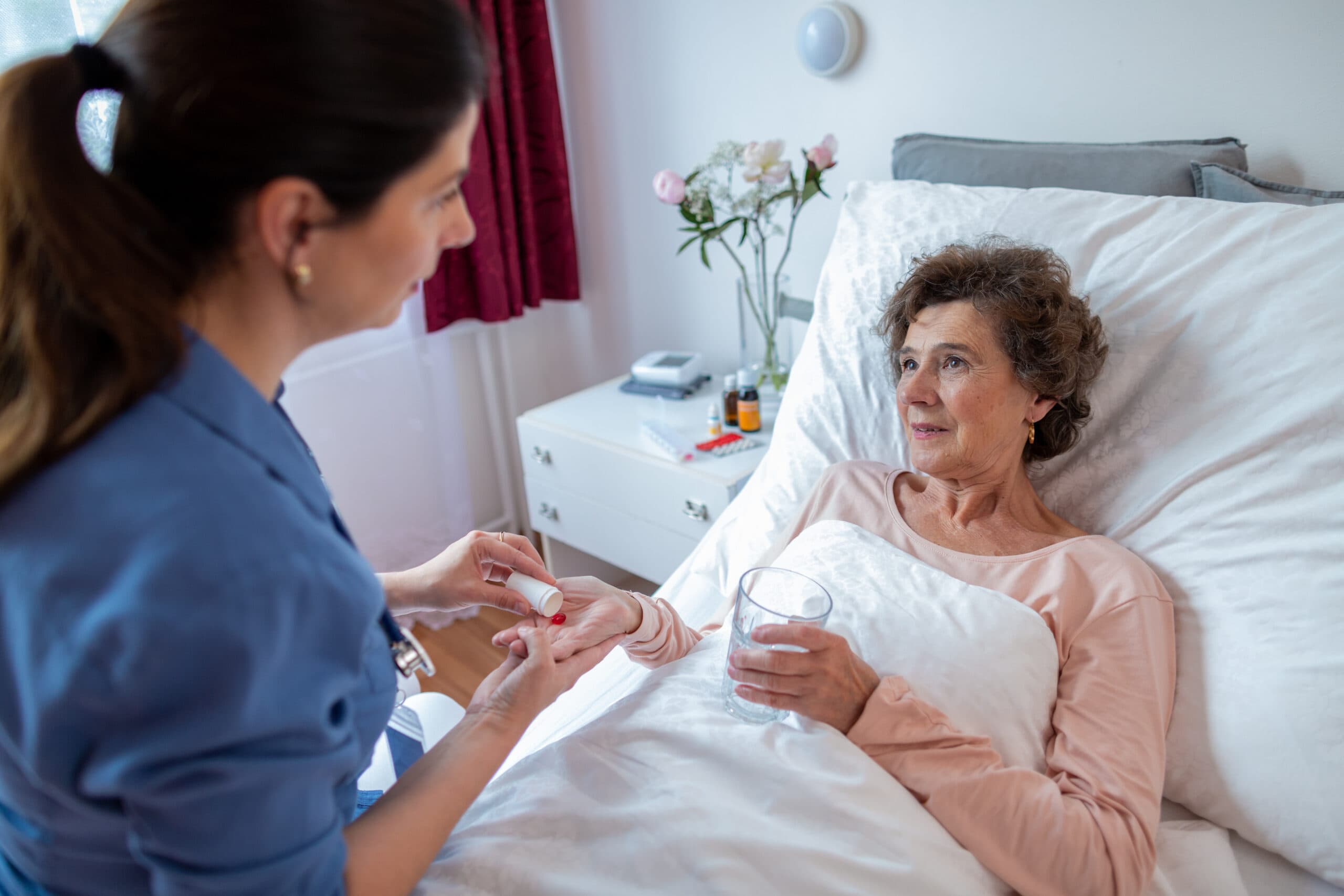 Elderly Female Patient Taking Pills from Home Caregiver. Nurse Giving Medication to Senior Woman Lying in Bed in Hospital. Common Misconceptions About 24 Hour Care